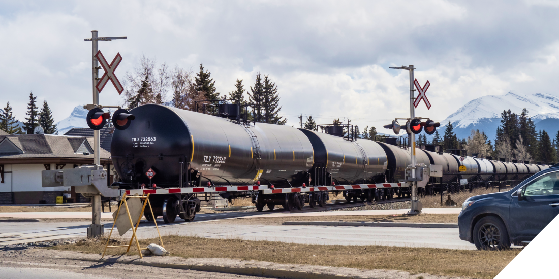 A black cylindrical tank car transporting oil, moving along railway tracks with mountains in the background.